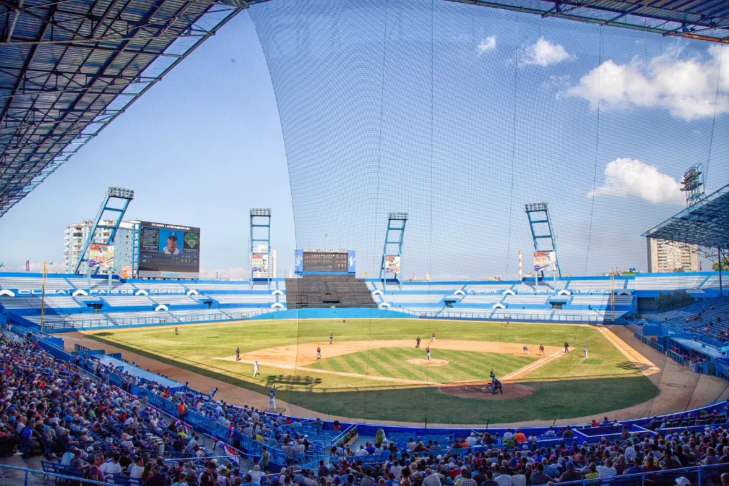 A wide view across a full Estadio Latinoamericano, blue seats packed, the green infield and floodlight towers under a bright sky.